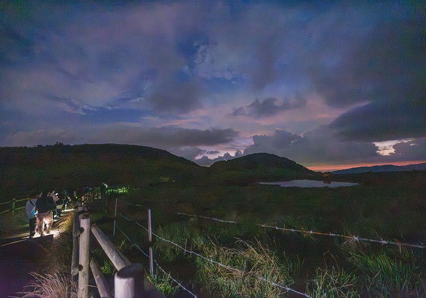 夜の牧野の中の通路から、遠くに火口跡の池が見える写真